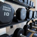Schwarze Hanteln mit Gewichtsangaben auf Regalen in einem Fitnessraum.