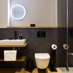 Badezimmer mit Waschtisch, Toilette und Dusche. Dunkle Wand, Holzregale und runder Spiegel.
