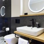 Badezimmer mit Dusche, Waschbecken und Toilette. Schwarze Wand, Holzoberfläche und beleuchtetes Spiegel.