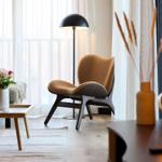 Ein Wohnbereich mit Sessel, Tisch und Stehlampe vor Fenster mit Vorhängen.