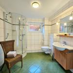 Bathroom with shower, sink, and toilet. Green tiled floor. Wicker chair with towels.