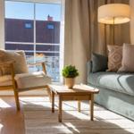 Wohnzimmer mit Sofa, Sessel, Tisch und Pflanze. Große Fenster mit Blick auf Balkon.