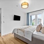 Schlafzimmer mit Bett, TV und Blick auf Balkon. Holzfußboden und blaue Vorhänge.