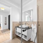 Badezimmer mit Dusche, Waschtisch und Spiegel. Blick in den Flur mit Holzboden.