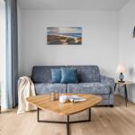 Ein Wohnzimmer mit grauem Sofa, Holztisch und Wandbild. Große Fenster mit Blick auf Gebäude.