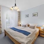 Schlafzimmer mit Holzbett, Nachttisch und Gardinen vor Fenster.