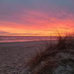 Sonnenuntergang über dem Strand mit Dünen und Wellen im Hintergrund.