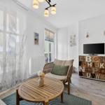 Wohnzimmer mit Sitzgelegenheit, TV und Holztisch. Fenster mit weißen Vorhängen.