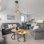 Wohnzimmer mit Sofa, Sessel und Essbereich. Hängelampe, Fenster und Küchenzeile sichtbar.