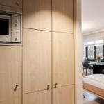Kitchen with integrated microwave and wooden cabinets. In the background, a bedroom with a bed and table.