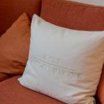 Weißer Kissenbezug mit geprägtem Ortsnamen und Koordinaten auf einem orangefarbenen Sofa.