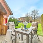 Terrasse mit Tisch, zwei Stühlen und Blick auf grünen Garten. Hintergrund: Haus mit Strohdach.