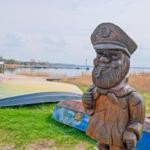 Holzfigur eines Matrosen mit Pfeife neben Bootsstegen am Wasser.