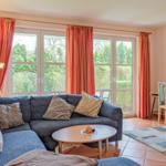 Ein Wohnzimmer mit Sofa, Tisch und Schreibtisch. Große Fenster mit orangefarbenen Vorhängen.
