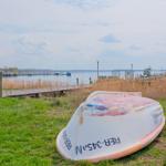 Eine rostige kleine Bootshülle liegt auf Gras nahe einem Steg am Wasser.