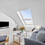 Wohnraum mit Dachfenster, Sofa, Fernseher und Tisch. Blick auf Strand.