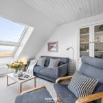 Dachgeschosswohnung mit großem Dachfenster, Sofa und Sessel. Blick auf Küstenlandschaft.