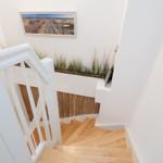 Helle Treppe mit Holzstufen und weißem Geländer. Wandbild und Pflanzen im Hintergrund.