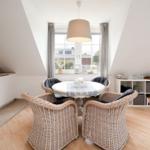 Esszimmer mit runden Tisch, vier Stühlen und großem Fenster. Weiße Küchenzeile und Holzboden.