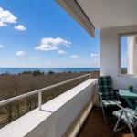 Balcony with table and chairs overlooking sea and forest under blue sky.
