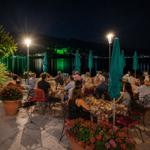 Guests sit at tables with flowerbeds and green umbrellas by the lakeside at night.