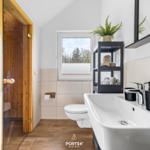 Badezimmer mit Dusche, Waschbecken und Toilette. Fenster zeigt Wald. Schwarze Regale und Pflanze.