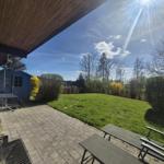 Terrace with grill, table and chairs, view of green lawn and trees under blue sky.