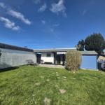 House with garden, sun loungers, and blue shed under blue sky.