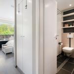Badezimmer mit Toilette und Regalen. Nebenraum mit Bett und Balkon.