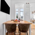 Essbereich mit Holztisch, braunen Stühlen und TV an der Wand. Blick in Wohnzimmer mit Sofa und Fenster.