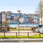 Uferpromenade mit Blick auf Häuser und Wasser. Parkplatz mit Autos und Bäumen.