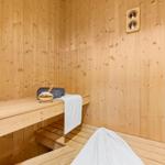 Sauna with wooden bench, towel, and basket. Wall clock and wooden walls visible.