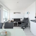 Living area with sofa, armchair, and kitchen. Large windows with sea view.