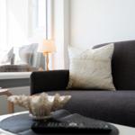 A dark sofa with a pillow, table with a shell and remote, window with decor.