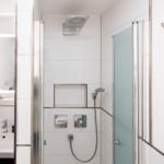 Shower with overhead and handheld shower, sink with mirror and small sailboat decoration.