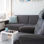 Ein grauer Sofa und Sessel stehen vor einem Fenster mit Tisch und Kissen.