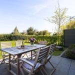 Terrace with table, chairs, and grill. Lawn and hedge in the background.