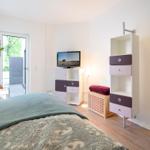 Bedroom with bed, TV, and cabinets. Door leads to balcony. Wooden floor and white walls.