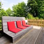 Wooden terrace with red cushions and wooden railing surrounded by greenery.