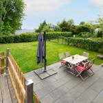 Terrace with table, chairs, and umbrella. Backyard with lawn and hedge.
