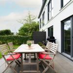 Deck with wooden table, red seat cushions, and grill beside the house.