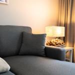 A gray sofa with cushions and a lamp on a table next to a window.
