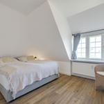 Schlafzimmer mit Bett, Nachttisch und Stuhl neben Fenster mit Vorhängen.