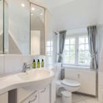 Badezimmer mit Waschbecken, Spiegel, Toilette und Fenster mit Vorhängen.