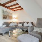 Wohnzimmer mit grauem Sofa, Sessel und Glas-Tisch unter Holzbalkendecke.