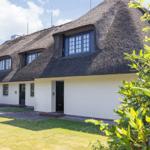 Haus mit strohgedecktem Dach, weißen Wänden und Fenstern mit dunklen Rahmen.