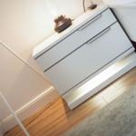 White nightstand with three drawers, lamp, and remote. Wooden floor and rug.