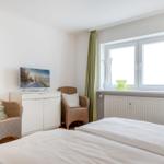 Bedroom with bed, two chairs, TV, and window with green curtains.
