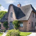 Ein Haus mit strohgedecktem Dach und Backsteinfassade. Gartenbereich mit Liegestühlen.
