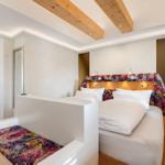 Schlafzimmer mit Bett, Kopfteil und Sitzbank mit floraler Tapete. Holzbalken an der Decke.